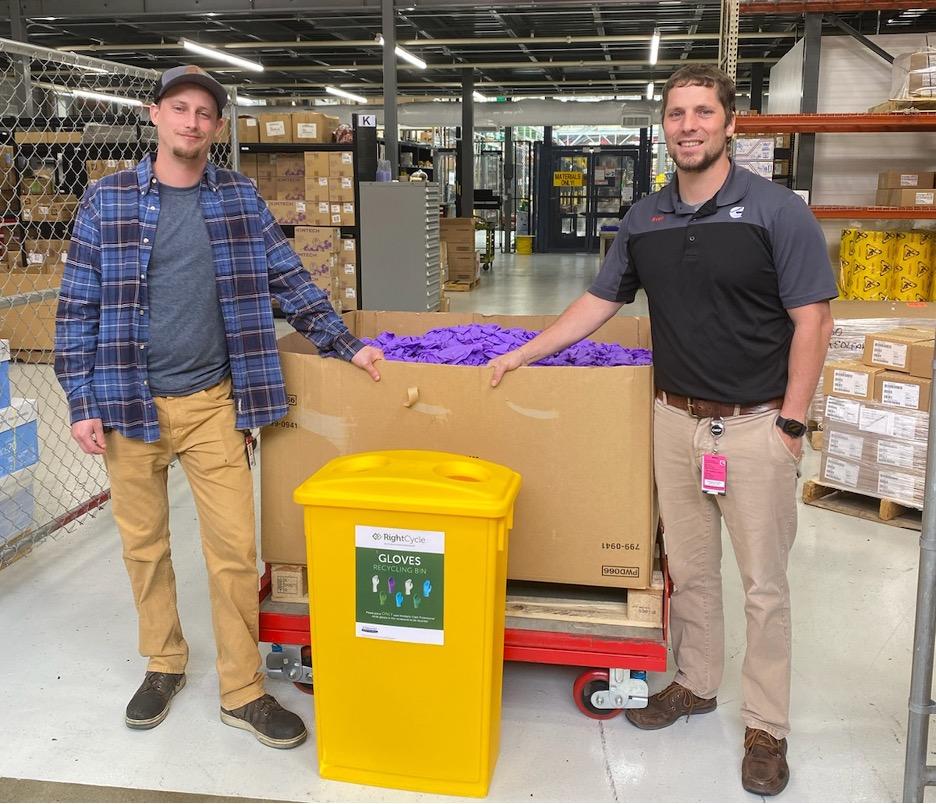 Continuous Improvement Leader, Clayton Johnson (left), and Materials Manager, Sven Creutz (right), stand next to a nitrile glove recycling container they implemented as part of the initiative. The receptacles are placed at every glove station in Columbus Mid-Range Engine Plant. ”“